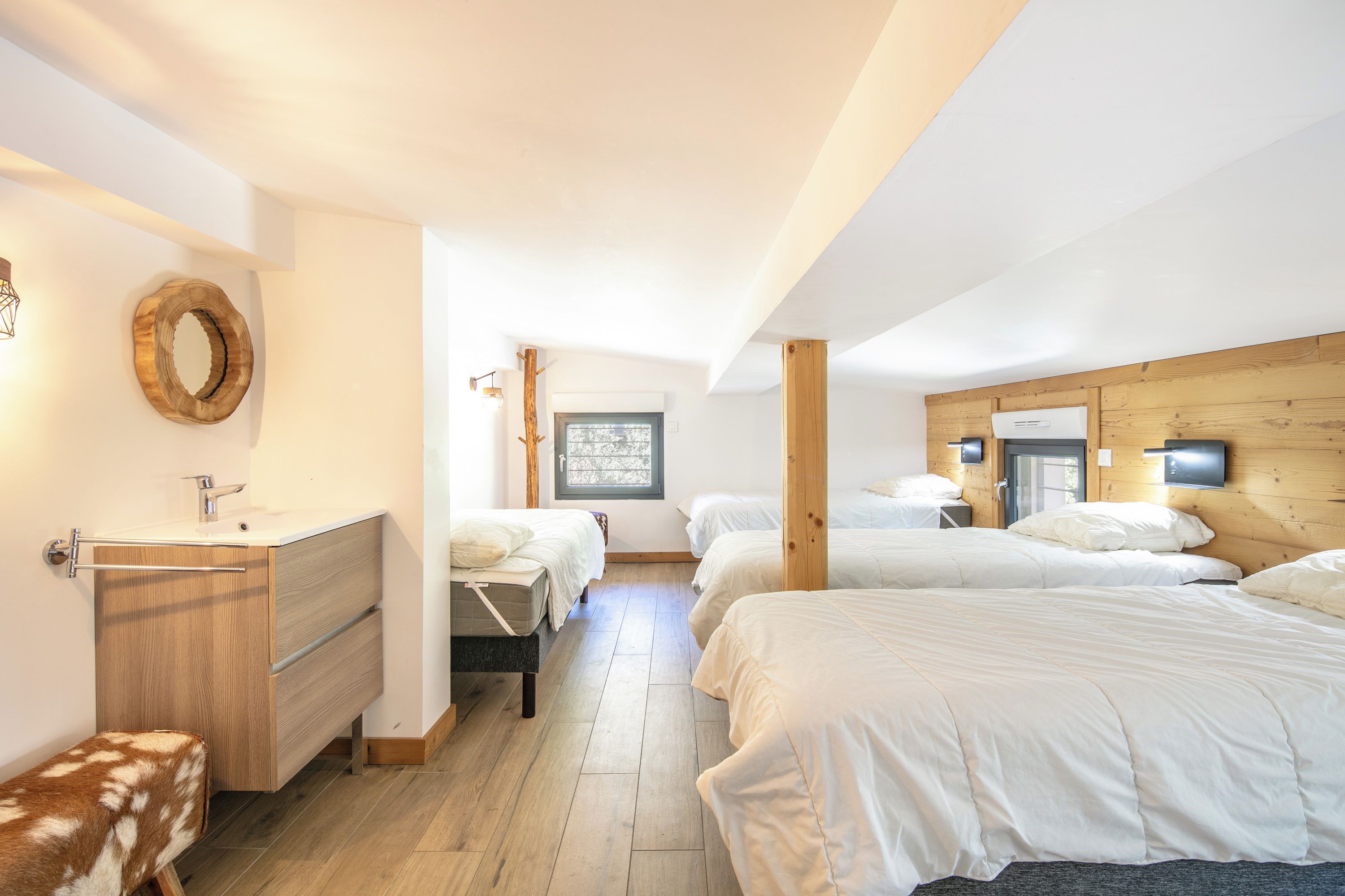 A bedroom with a wooden headboard, a wooden bench, a round mirror, a bedside table, and a window.