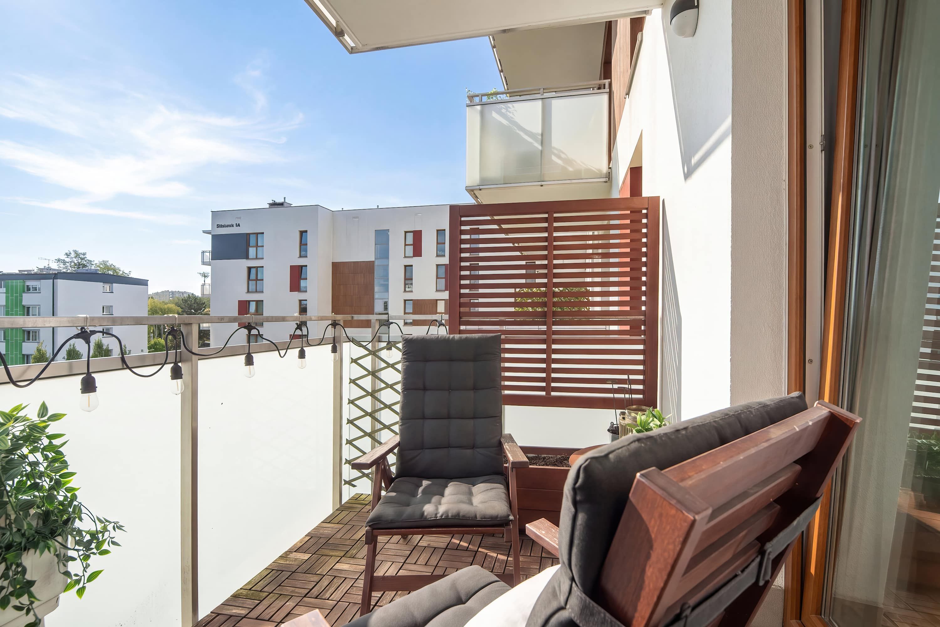A balcony with a wooden chair and a small table, a potted plant, and a view of a building with multiple windows.