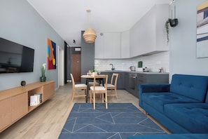 Een moderne keuken met witte kasten, een eethoek met tafel en stoelen, een blauwe bank en een aan de muur bevestigde televisie.