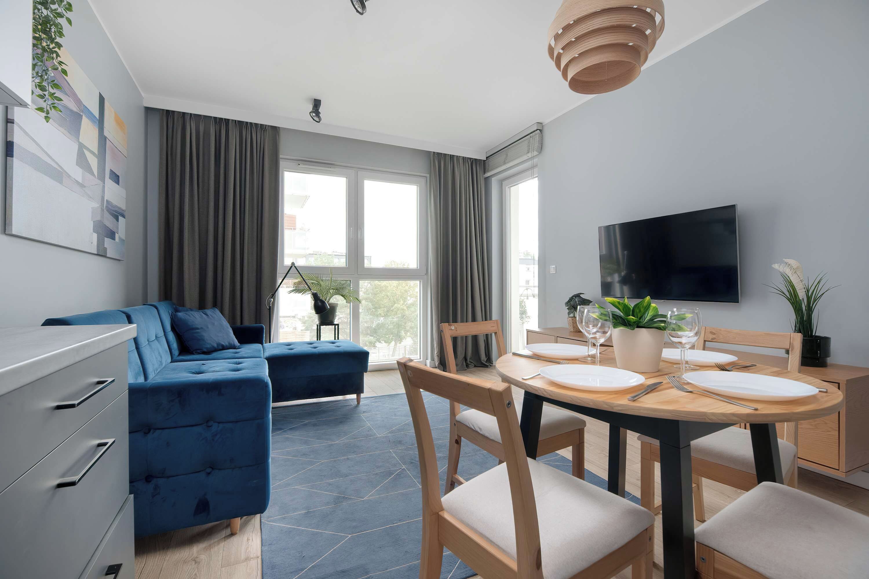 Uma sala de estar moderna com um sofá azul, uma mesa de jantar posta para quatro pessoas e uma TV fixada na parede.