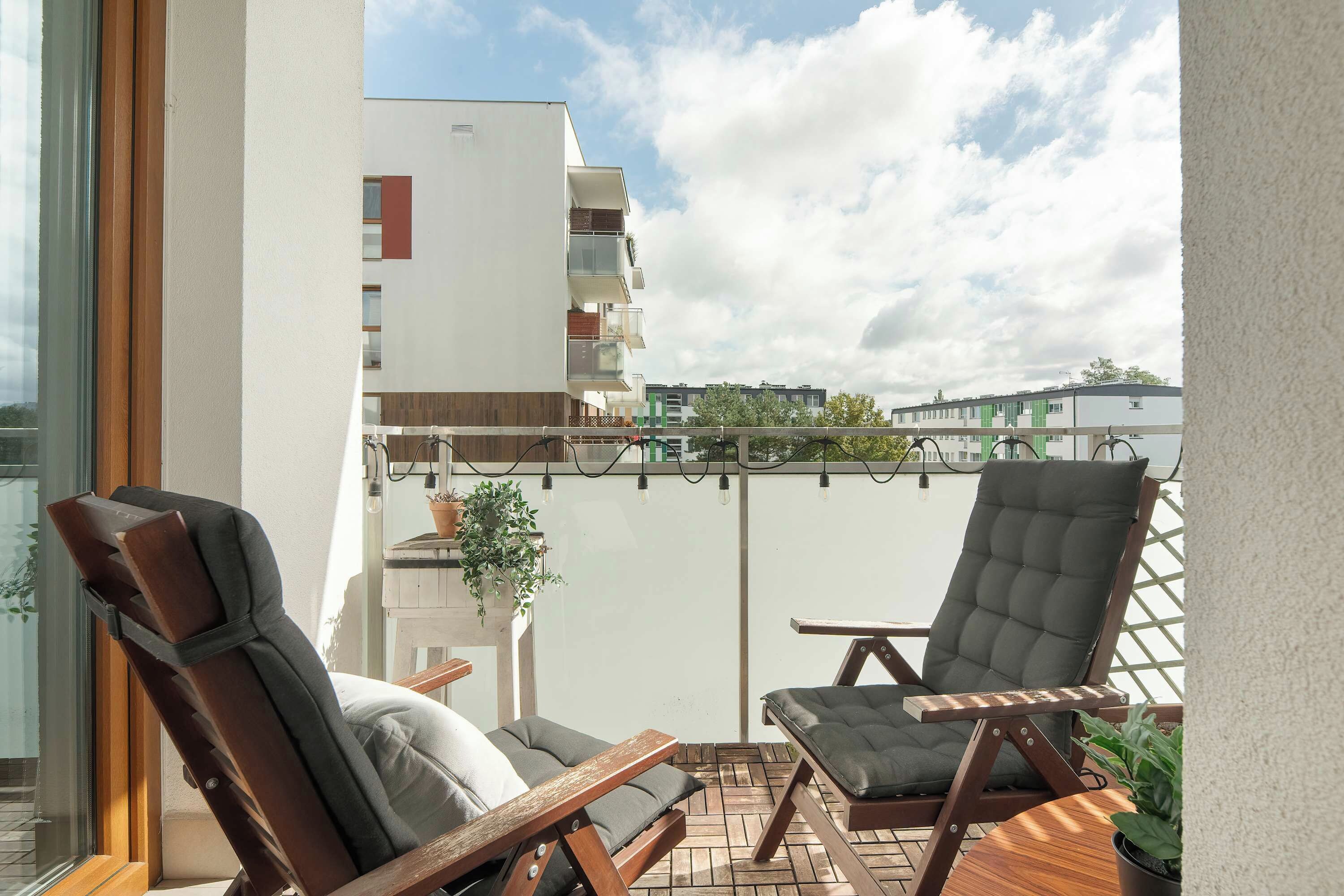 Un balcon doté de deux fauteuils de jardin en bois et d’une petite table, offrant une vue sur un quartier résidentiel avec des immeubles modernes.