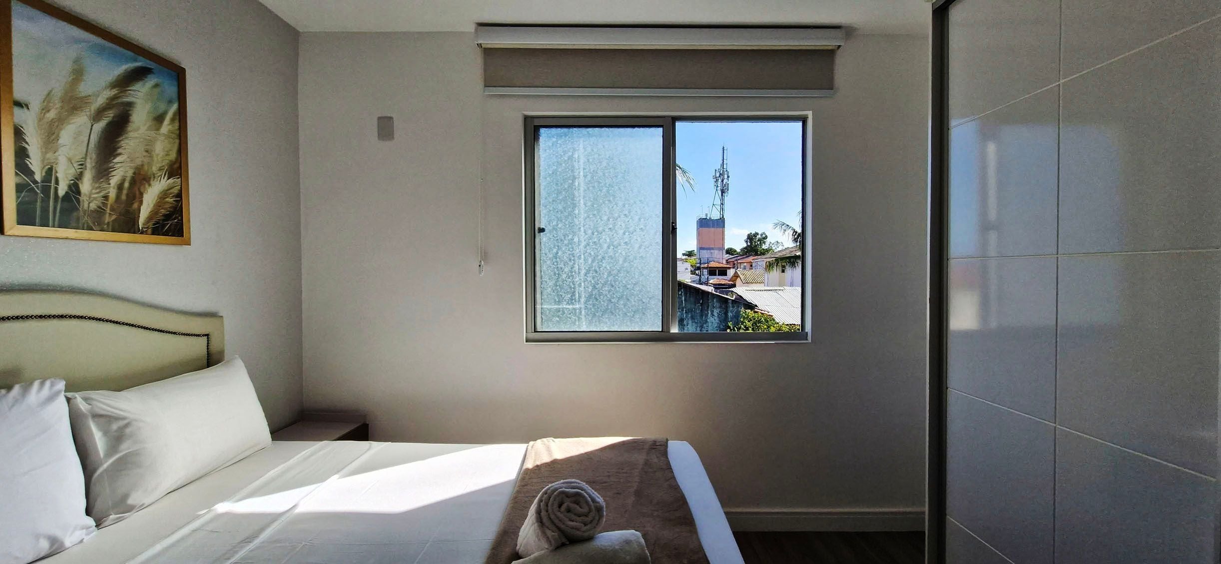 A hotel room with a bed, a window with a view of buildings and a communication tower, and a painting on the wall.