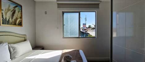 A hotel room with a bed, a window with a view of buildings and a communication tower, and a painting on the wall.