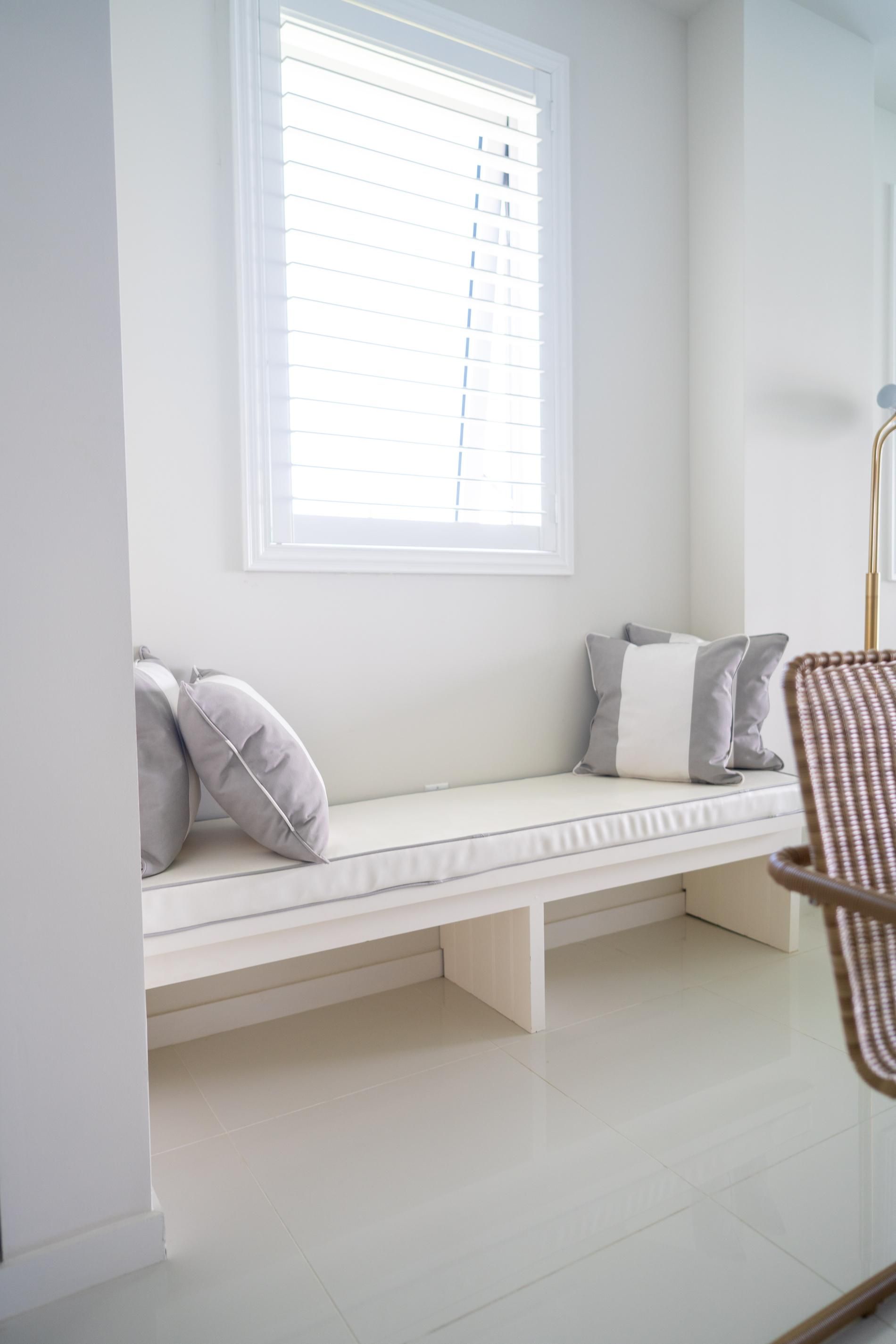 Une pièce avec un banc blanc adossé à un mur, une fenêtre avec des stores et une chaise dont l’assise est en osier tressé.