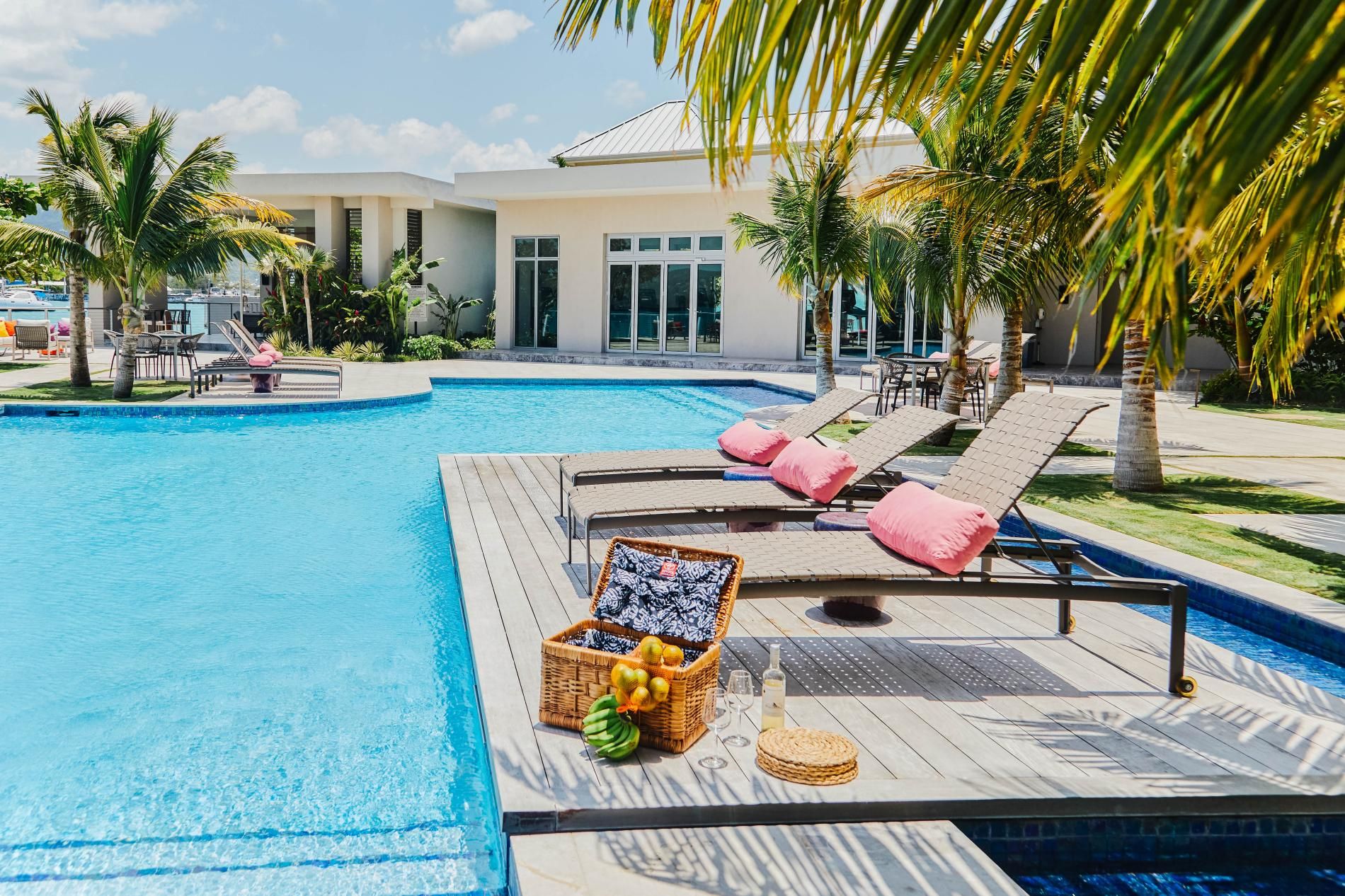 A swimming pool with lounge chairs and a wicker basket containing fruits and a bottle.