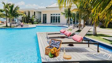 A swimming pool with lounge chairs and a wicker basket containing fruits and a bottle.