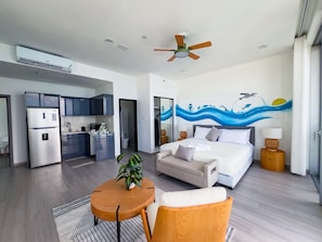 A modern hotel room with a bed, a sofa, a dining table, and a kitchen area.