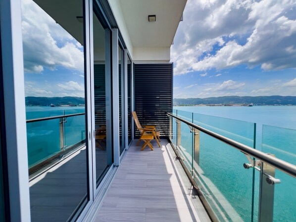 A balcony with a glass railing, wooden deck, and a view of the sea and mountains.