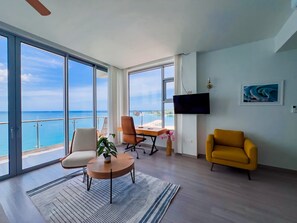 A spacious living area with a large window overlooking the sea, a flat-screen TV mounted on the wall, and a yellow armchair.
