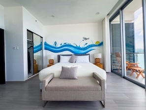 Ein modernes Schlafzimmer mit einem großen Bett, Meerblick durch eine Schiebetür und ein Balkon mit Sitzmöbeln.