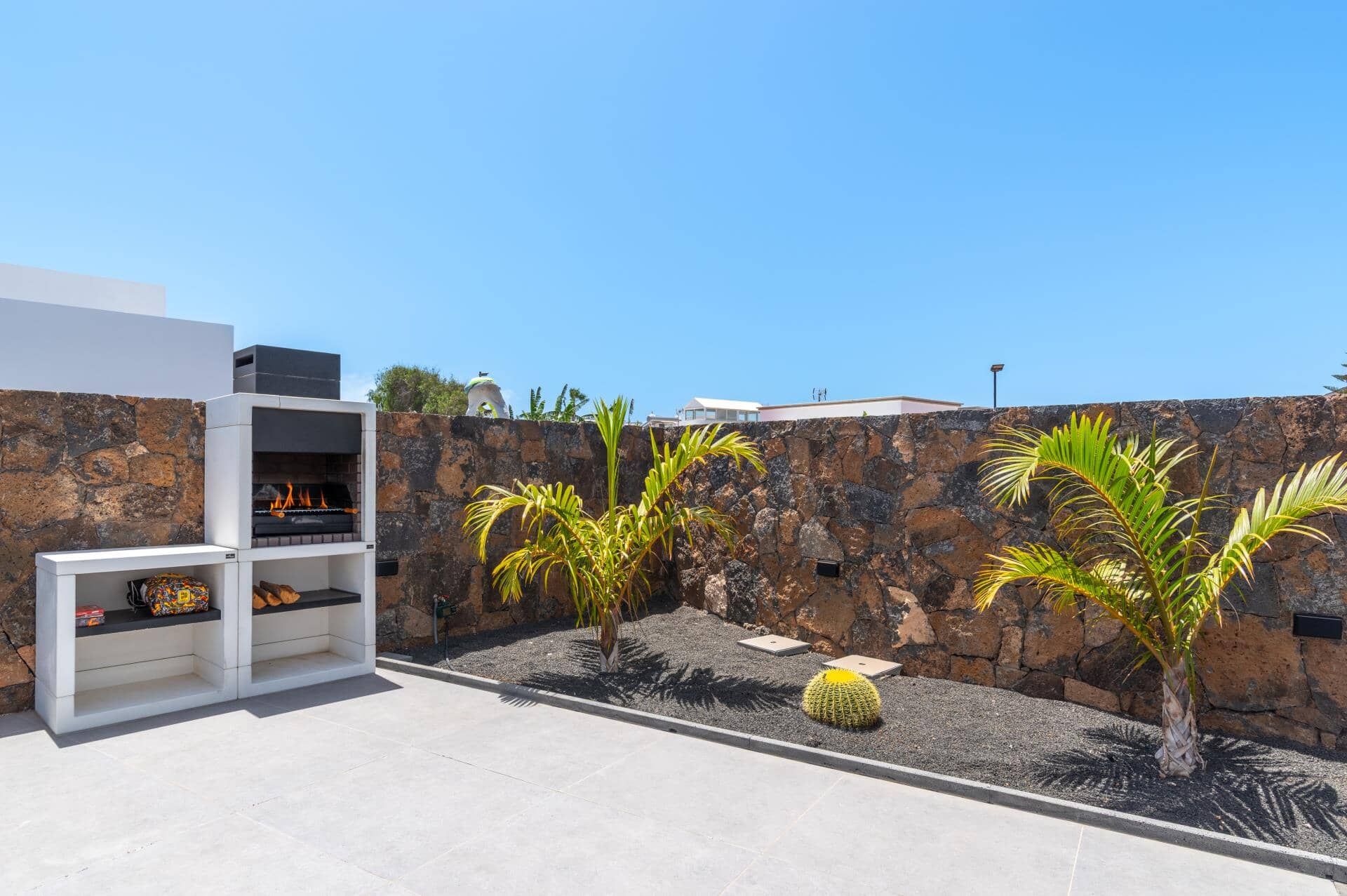 A modern outdoor patio with a built-in barbecue grill, palm trees, and a stone wall.