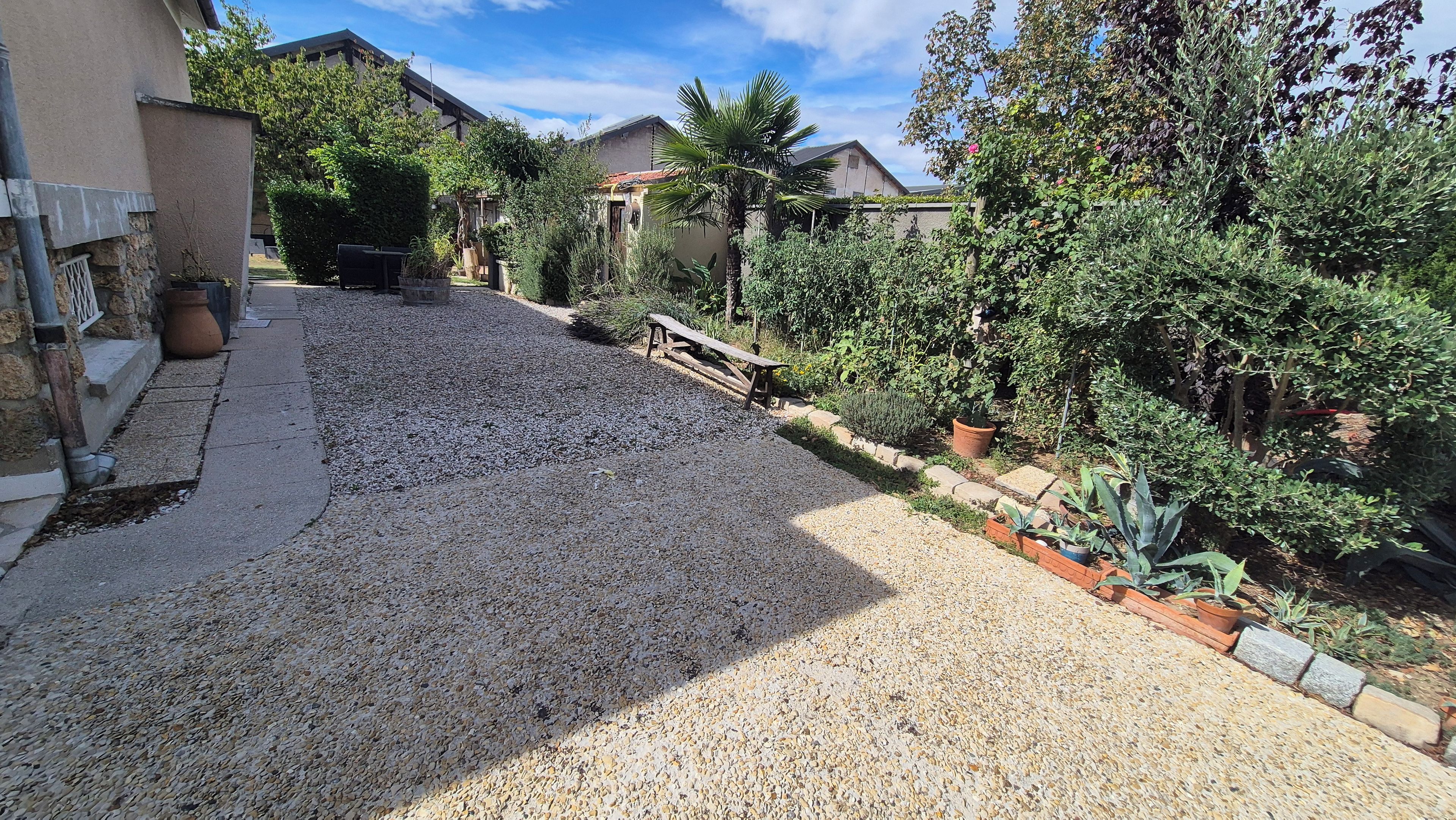 Ein Kiesweg führt zu einem Haus mit Garten, einer Holzbank und einem Blumentopf.