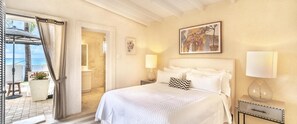 A hotel room with a bed, a nightstand, a lamp, a painting, and a view of the ocean through a door. - Little Good Harbour House- 3BR Ocean Front Villa (St. Peter)