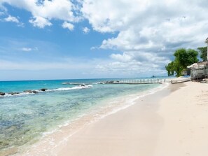 A beach with clear water, a wooden pier, and a building with a balcony. - Little Good Harbour House- 3BR Ocean Front Villa (St. Peter)