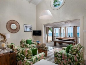A living room with a sofa set, a coffee table, a TV, and a view of the ocean through large windows. - Little Good Harbour House- 3BR Ocean Front Villa (St. Peter)