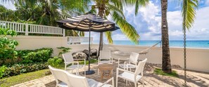 Een terras met witte stoelen en een tafel, een gestreept parasol en uitzicht op de oceaan.