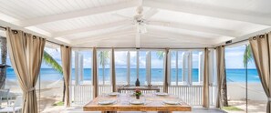 Uma área de refeições coberta com vista para o oceano, uma mesa de madeira posta para refeição e um ventilador de teto.