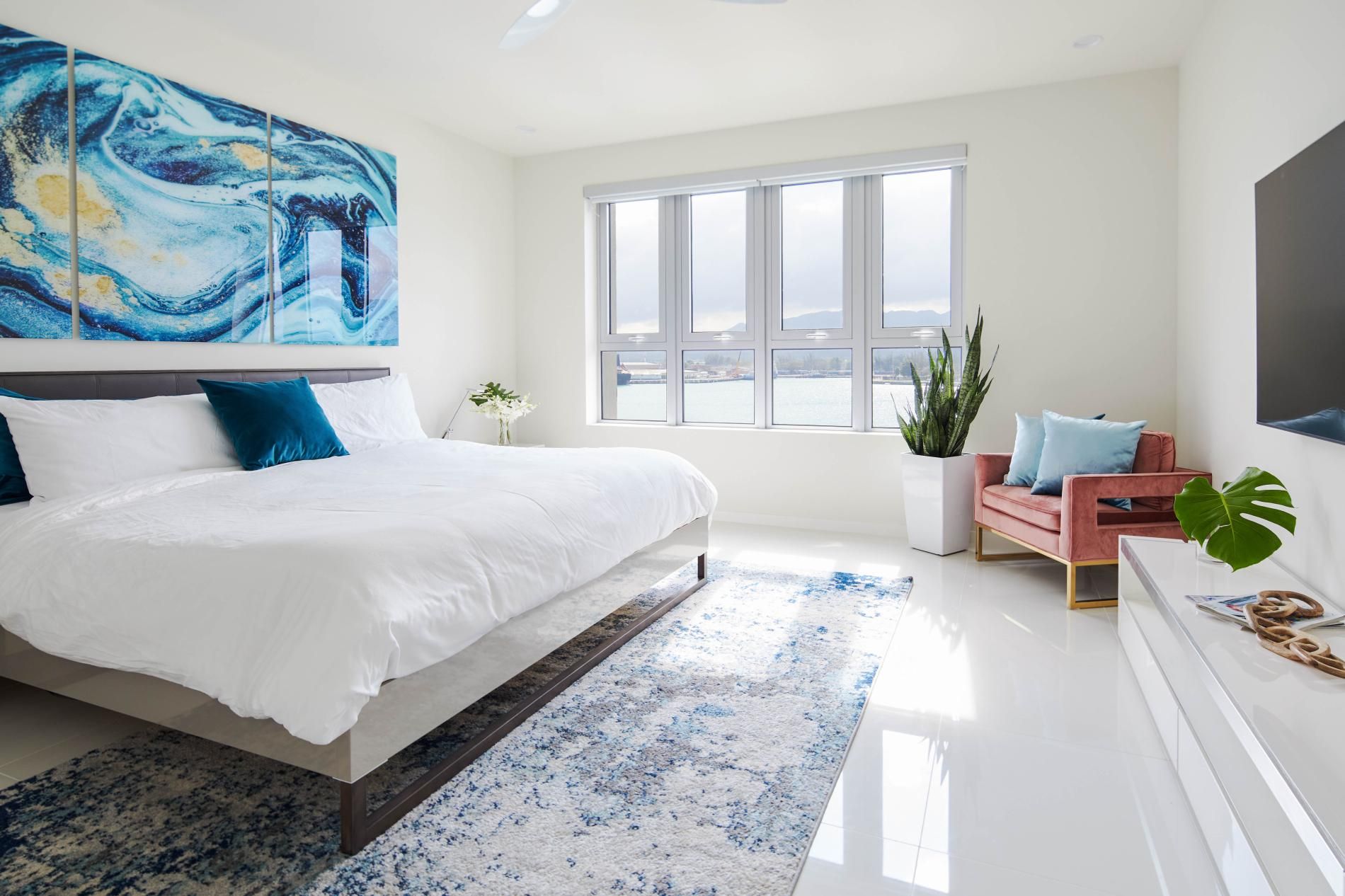 Ein modernes Schlafzimmer mit einem großen Bett, einem Fenster mit Stadtblick und einem blauen, abstrakten Gemälde.