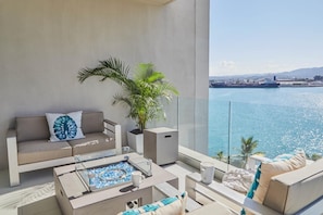 A balcony with a sofa, a coffee table, and a potted plant, overlooking a body of water and a distant ship. - Live Life-3BR Luxury Oceanfront Condo in Freeport (Montego Bay)