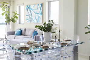 A modern living room with a glass dining table, clear chairs, a grey sofa with blue cushions, and two large abstract paintings on the wall. - Live Life-3BR Luxury Oceanfront Condo in Freeport (Montego Bay)