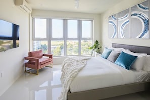 Une chambre moderne avec un grand lit, un fauteuil rouge et une vue sur la ville à travers de grandes fenêtres.