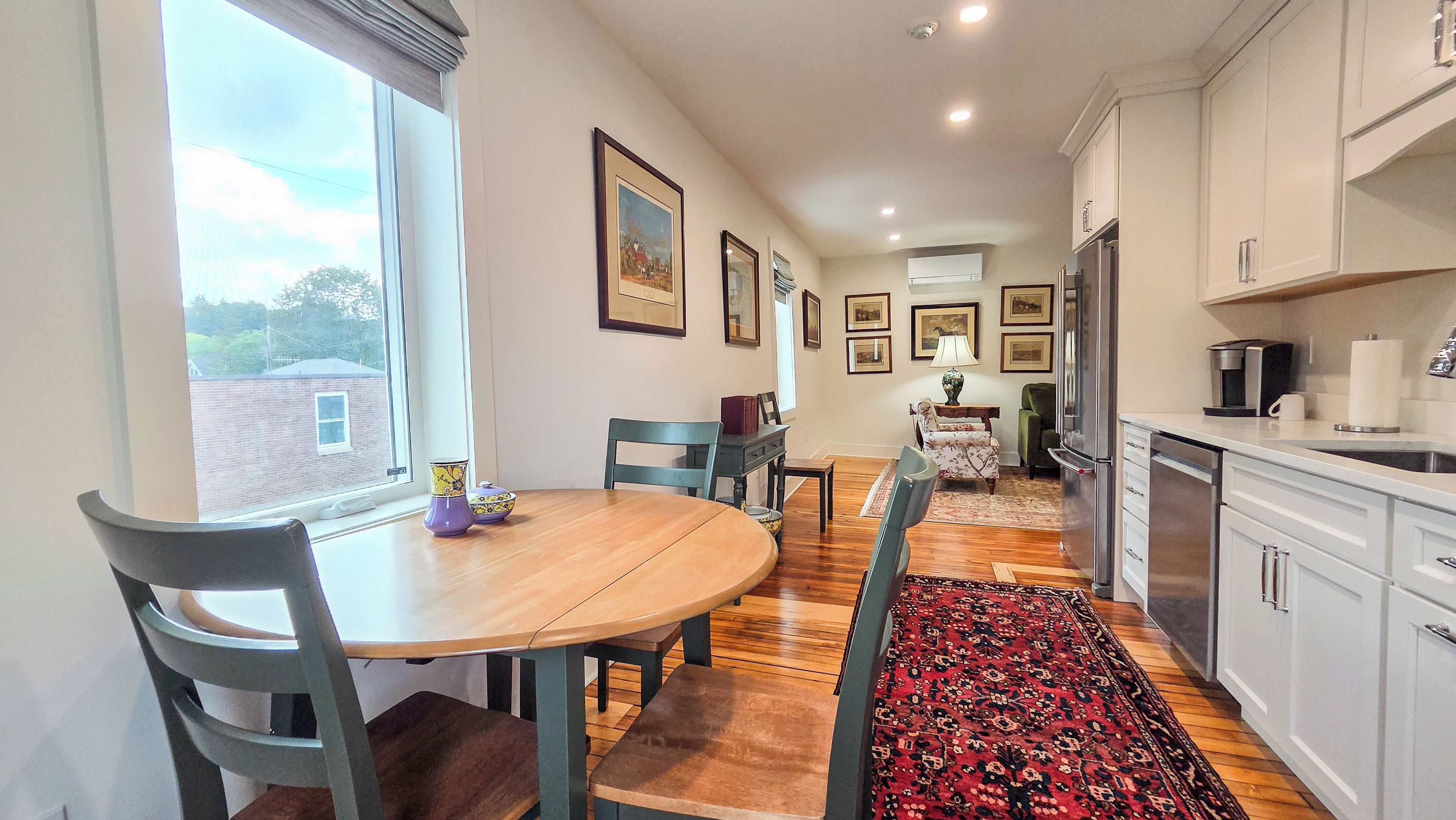 A dining area with a wooden table, chairs, and a rug. There are framed pictures on the wall, a window with curtains, and a kitchen area with cabinets and appliances.