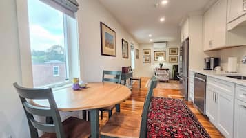A dining area with a wooden table, chairs, and a rug. There are framed pictures on the wall, a window with curtains, and a kitchen area with cabinets and appliances.
