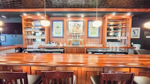 Angolo bar con bancone in legno, sgabelli e scaffali pieni di bicchieri e bottiglie.
