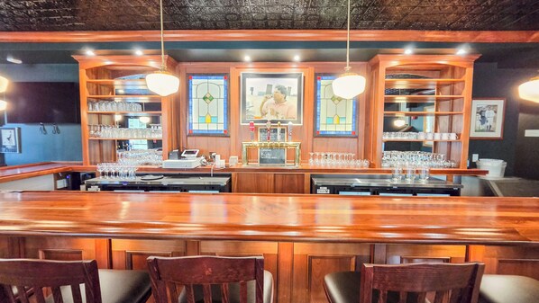 Zona de bar con mostrador de madera, varios taburetes y estantes con copas y botellas.