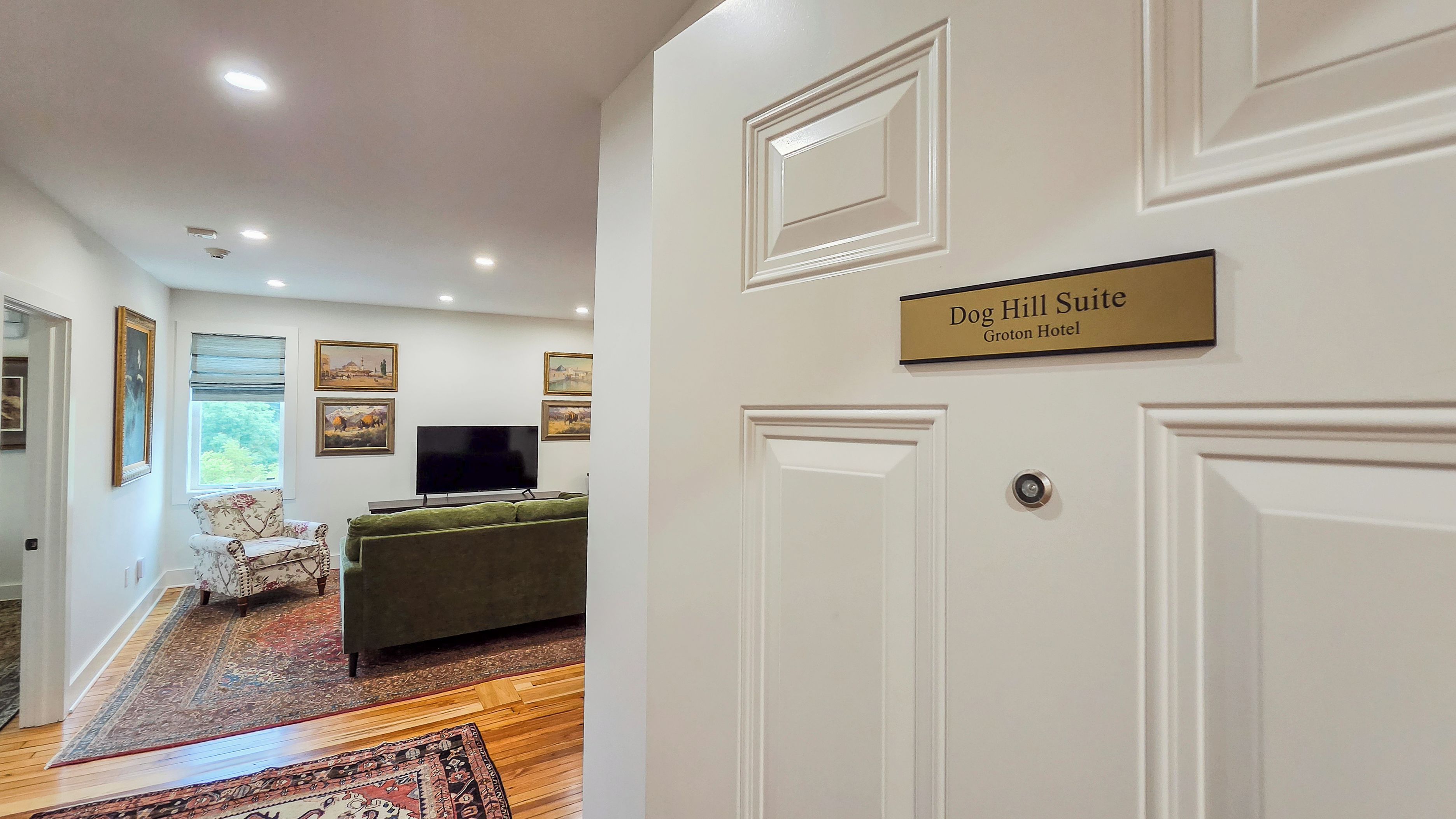 A hotel suite with a flat-screen TV, a sofa, and a dining table. There are framed pictures on the wall and a window with a view.