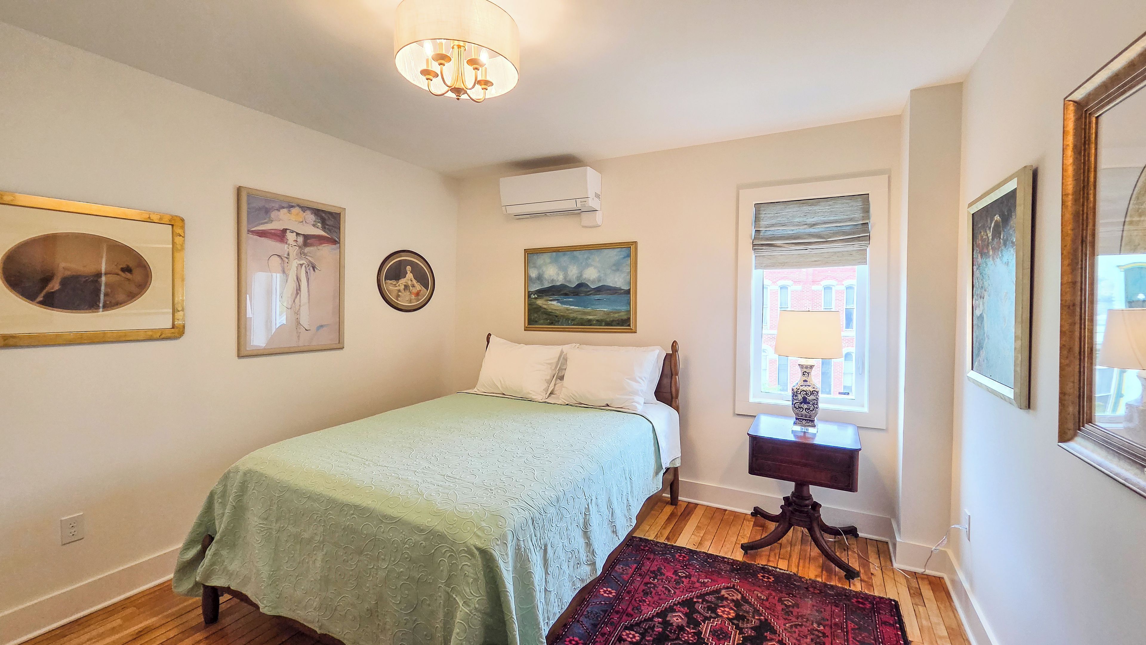 A bedroom with a bed, a window with a view, a lamp, and artwork on the walls.