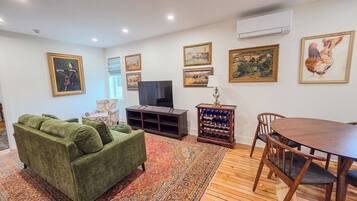 A living room with a green sofa, a TV, a wooden table with chairs, and a wine rack.