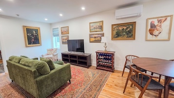 Una sala de estar con un sofá verde, un televisor, una mesa de madera con sillas y una estantería para vinos.