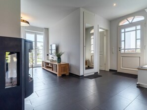 Ein geräumiger Wohnbereich mit dunklem Fliesenboden, einem Holzkonsolentisch mit Spiegel und Pflanze sowie einem großen Fenster, das natürliches Licht hereinlässt.