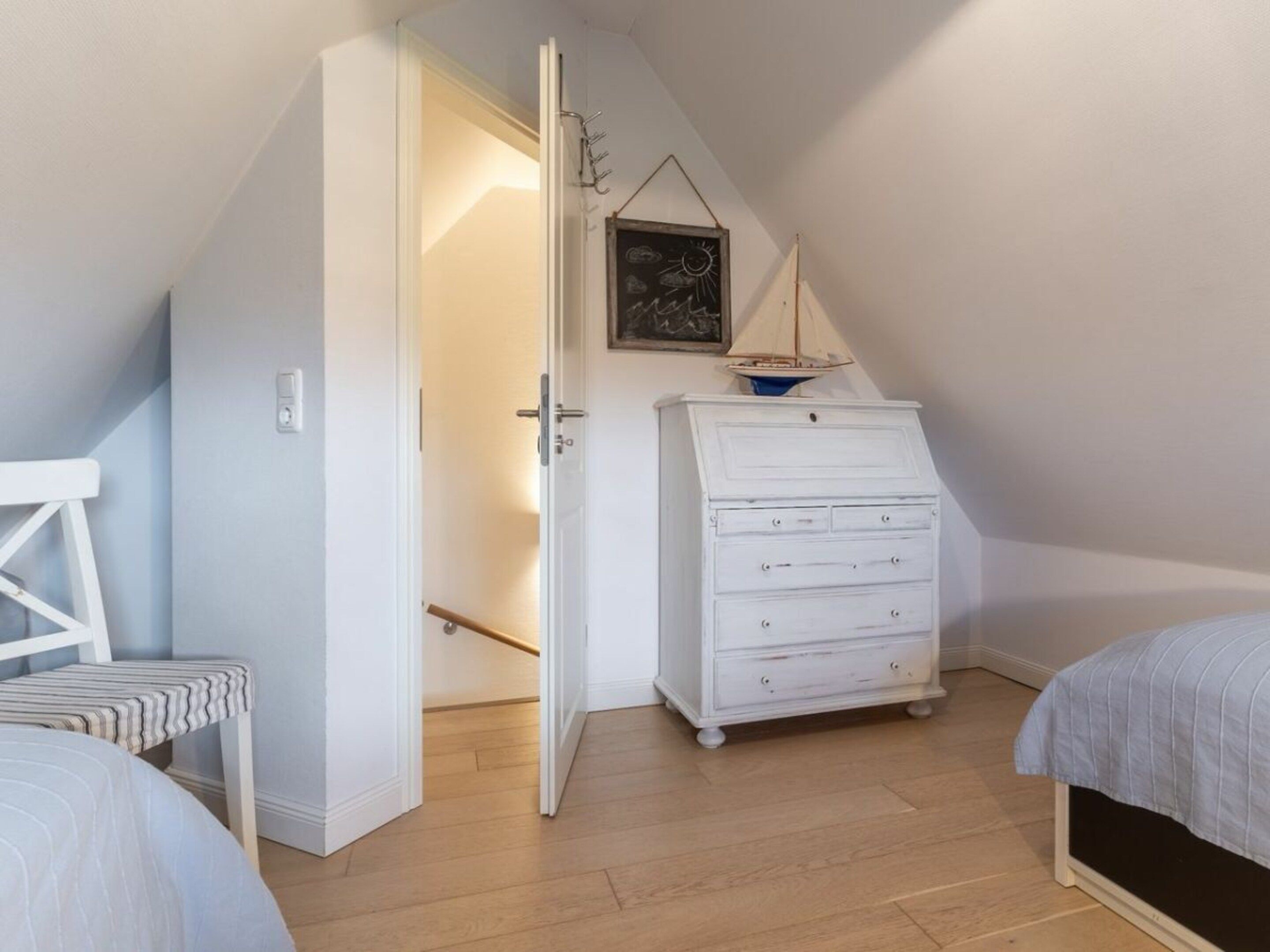 Ein Schlafzimmer mit schrägem Dach, einer weißen Kommode, einem Bett und einer kleinen Segelschiff-Dekoration.