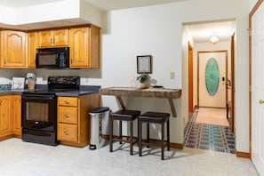 Une cuisine compacte avec des meubles en bois, un four à micro-ondes et une cuisinière. On y trouve un petit coin bar avec des tabourets et une poubelle. Un tableau encadré est accroché au mur.