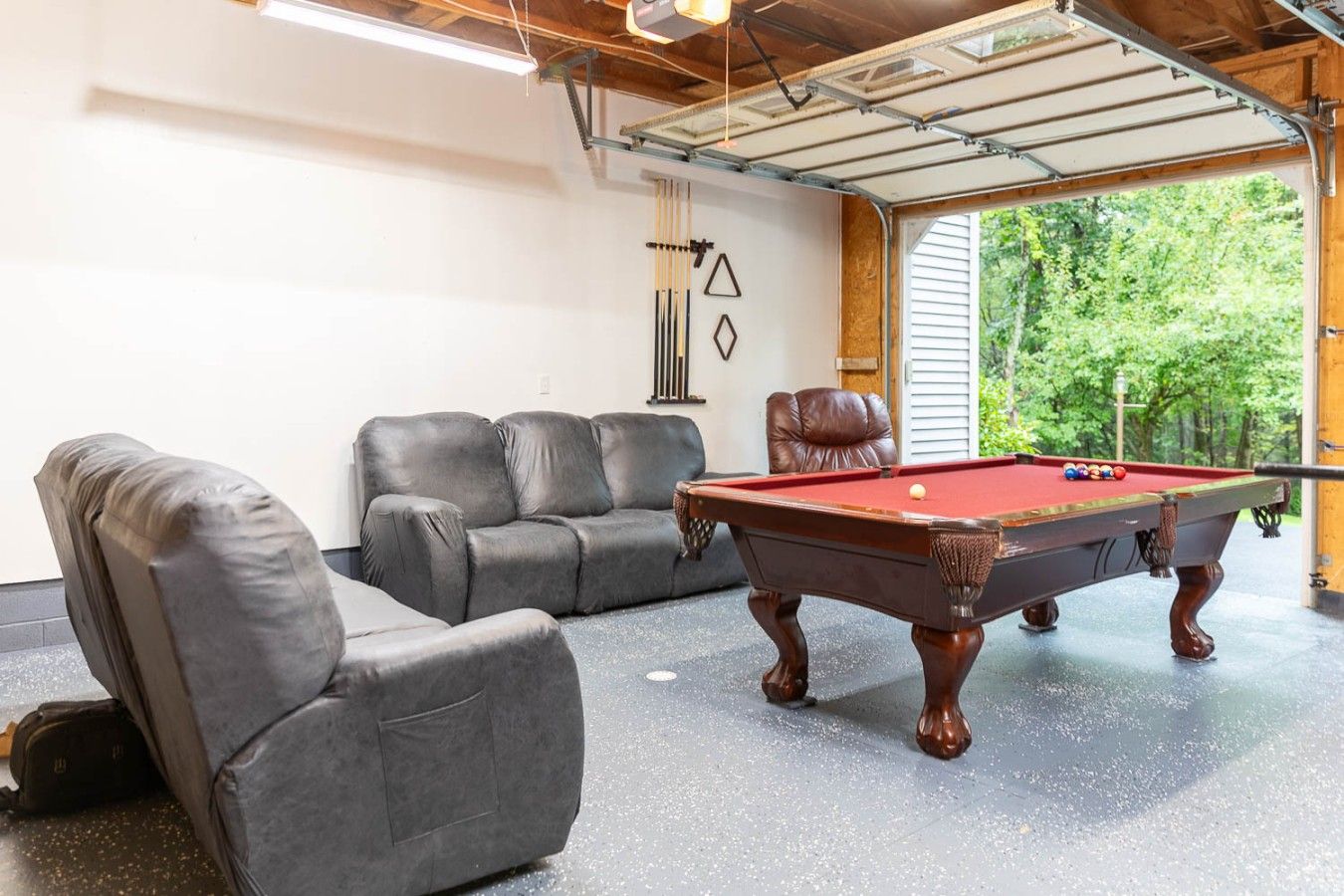 Une table de billard dans une pièce dotée d’une porte de garage, de deux fauteuils en cuir et offrant une vue sur la verdure à l’extérieur.