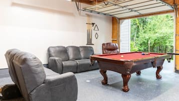 Une table de billard dans une pièce dotée d’une porte de garage, de deux fauteuils en cuir et offrant une vue sur la verdure à l’extérieur.