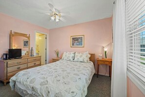 Ein Schlafzimmer mit einem Bett, einem Schminktisch, einem Fernseher, einem Deckenventilator, einem Nachttisch mit Lampe und einem Fenster mit Jalousien.