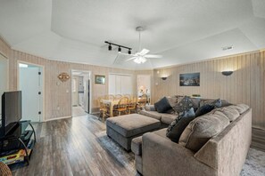 A living room with a sofa, ottoman, and dining area with chairs. - Guest Cottage 61- 4BR House- Community Pool (Myrtle Beach)