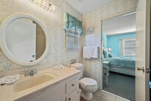 Ein Badezimmer mit Waschbecken, Spiegel, Toilette und einer Tür zu einem Schlafzimmer mit Bett und Fenster.