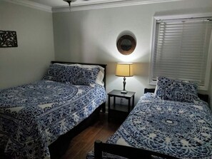 Ein Zimmer mit zwei Betten, einem kleinen Tisch mit Lampe, einem runden Spiegel an der Wand und einem Fenster mit Jalousien.