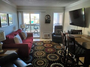 Un salon avec un canapé rouge, un tapis à motifs, un téléviseur et un balcon aménagé avec du mobilier d’extérieur.
