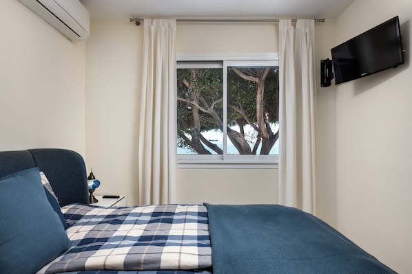 A bedroom with a bed, a TV mounted on the wall, a window with curtains, and an air conditioning unit.