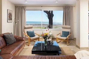 A living room with a view of the beach, two armchairs, a coffee table, and a brown leather sofa.