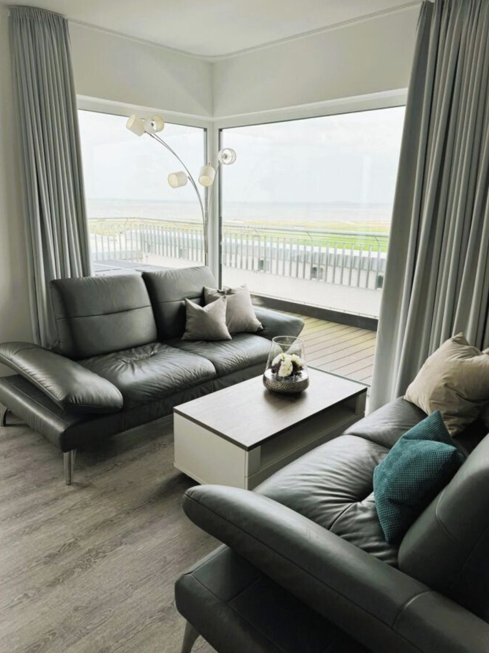 Een moderne woonkamer met een grijze leren bank, een witte salontafel en uitzicht op zee door grote ramen.