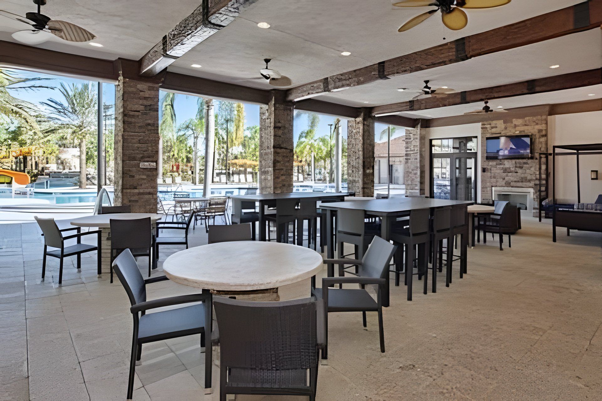 A spacious outdoor dining area with multiple tables and chairs, a pool in the background, and a stone wall.