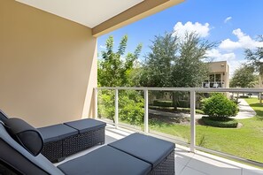 Ein Balkon mit zwei Loungestühlen, einer Glasreling und Blick auf eine grüne Landschaft mit Bäumen und einem Gebäude im Hintergrund.