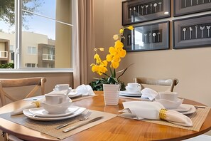 Ein Esstisch gedeckt mit weißem Geschirr, eine Vase mit gelben Blumen und gerahmtes Besteck an der Wand.