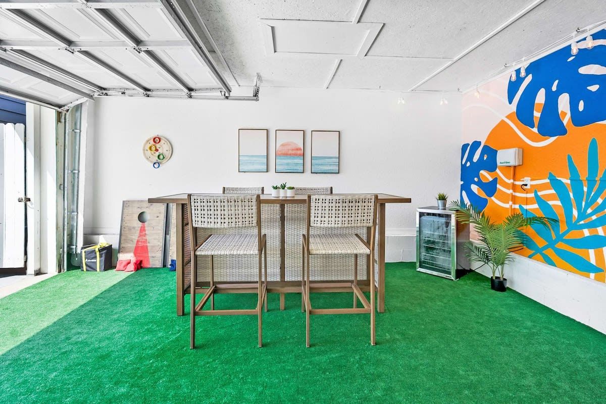 A room with a green artificial turf floor, a wooden table with two wicker chairs, and a wall with abstract art and a circular light fixture.